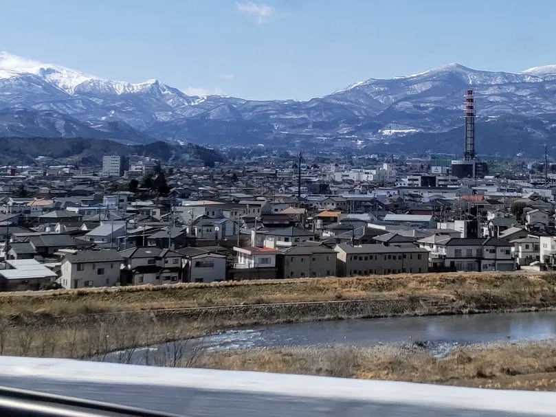 View from the Shinkansen of a river and town in Fukushima, with majestic snow-covered mountains towering in the background